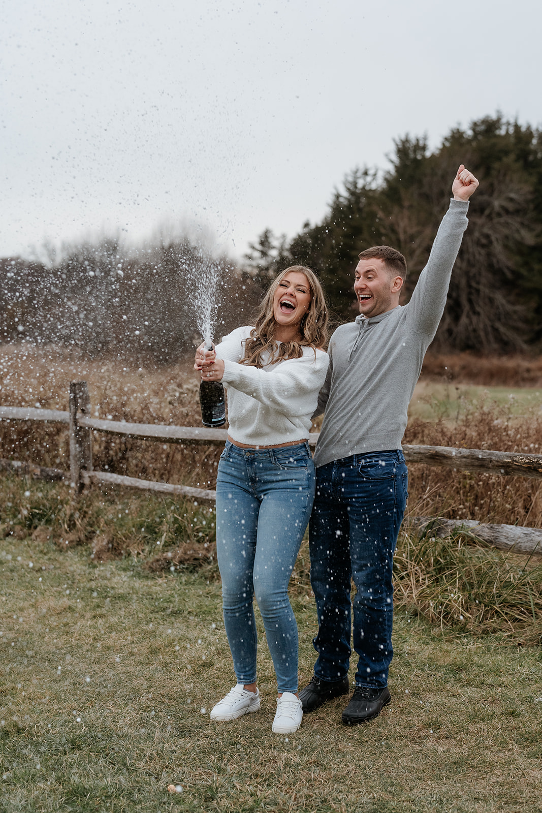 A couple popping champagne in a field during their engagement session.