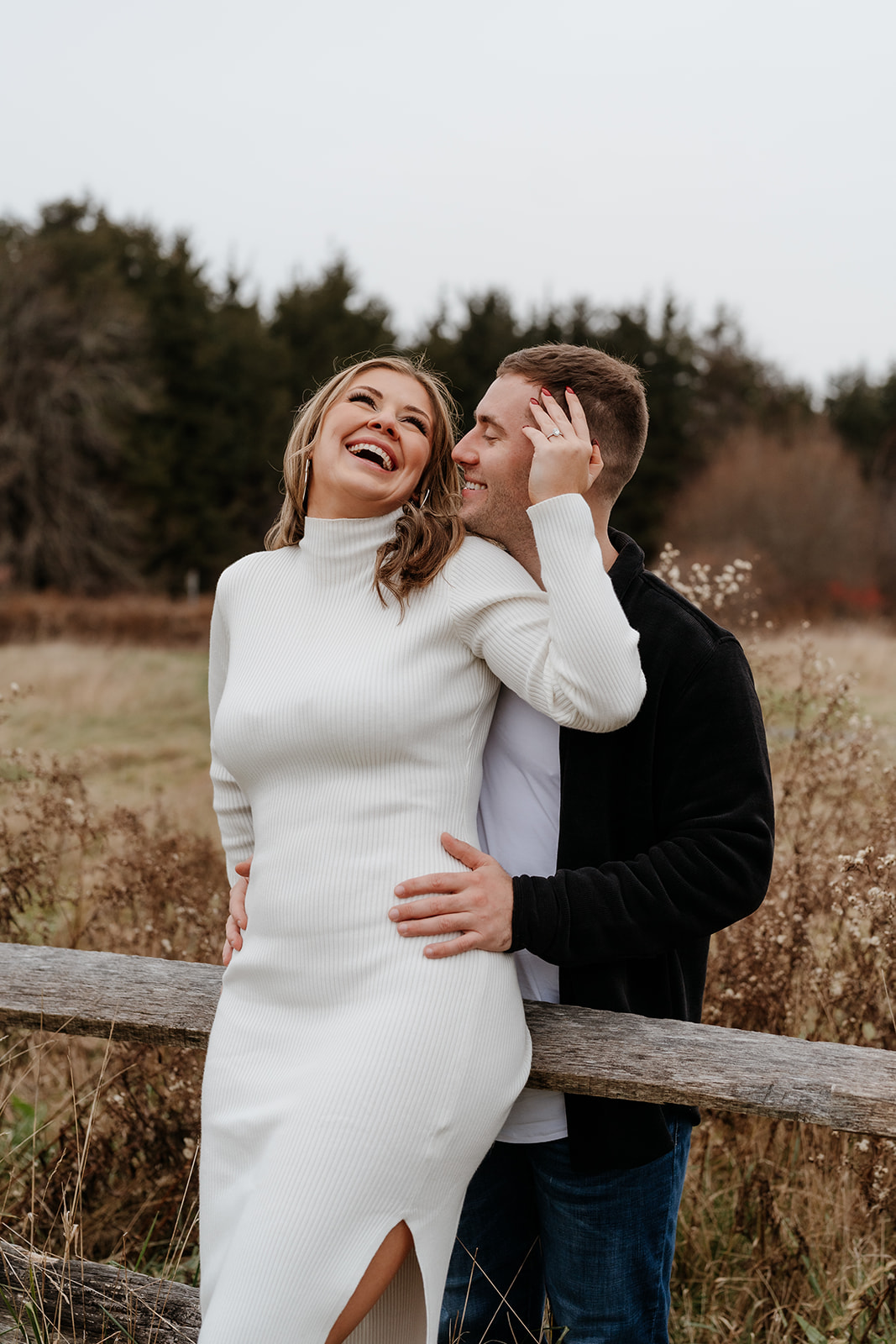 An engaged couple in a white dress leaning against a fence in a field during their documentary engagement session.