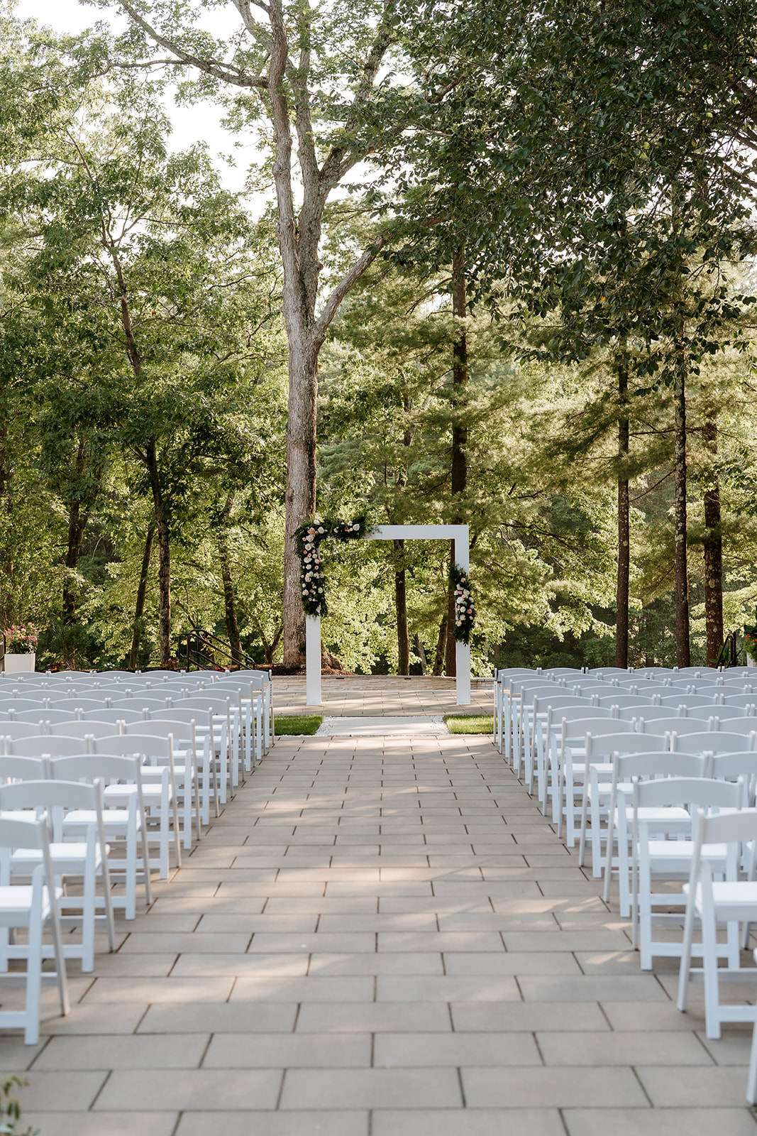 An outdoor wedding ceremony setup with white chairs aligned on either side of a stone aisle leading to a simple white arch adorned with floral arrangements, surrounded by green trees at lakeview pavilion.