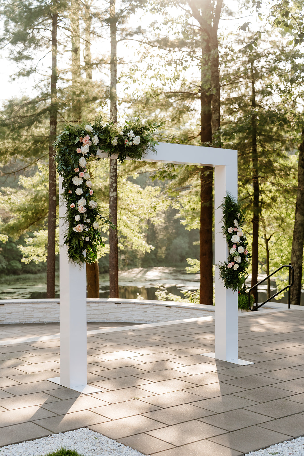 An elegant white ceremonial arch decorated with floral arrangements stands on a paved area surrounded by tall trees bathed in sunlight at Lakeview Pavilion