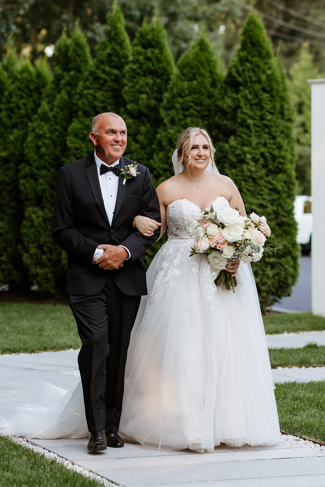 A bride in a white wedding gown holding a bouquet walks arm in arm with an older man in a black tuxedo, both smiling as they proceed along a pathway bordered by conical shrubs.