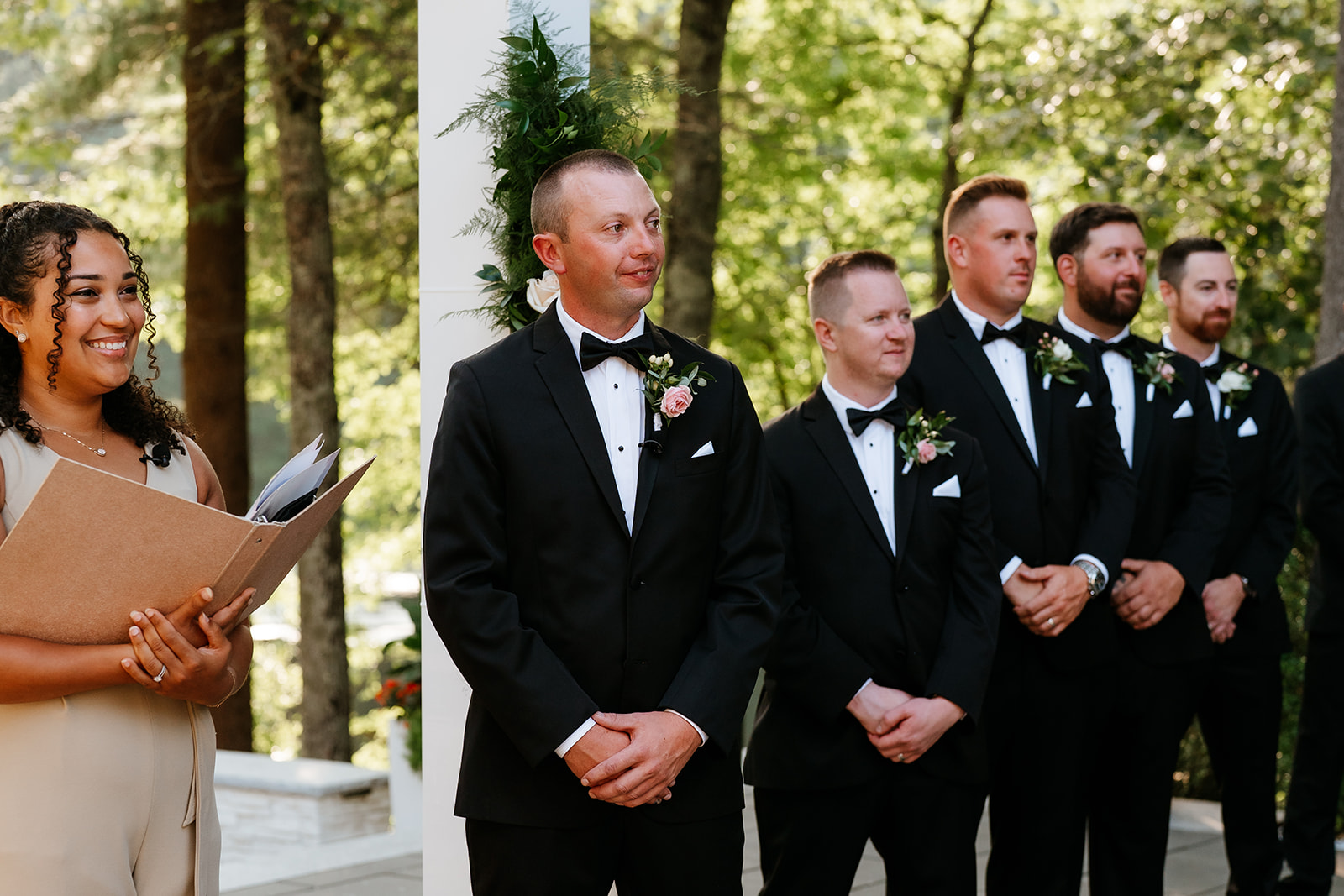 A group of groomsmen stands in a line wearing black tuxedos with boutonnieres, attentively watching an event, alongside a woman holding a folder, possibly officiating or coordinating,.