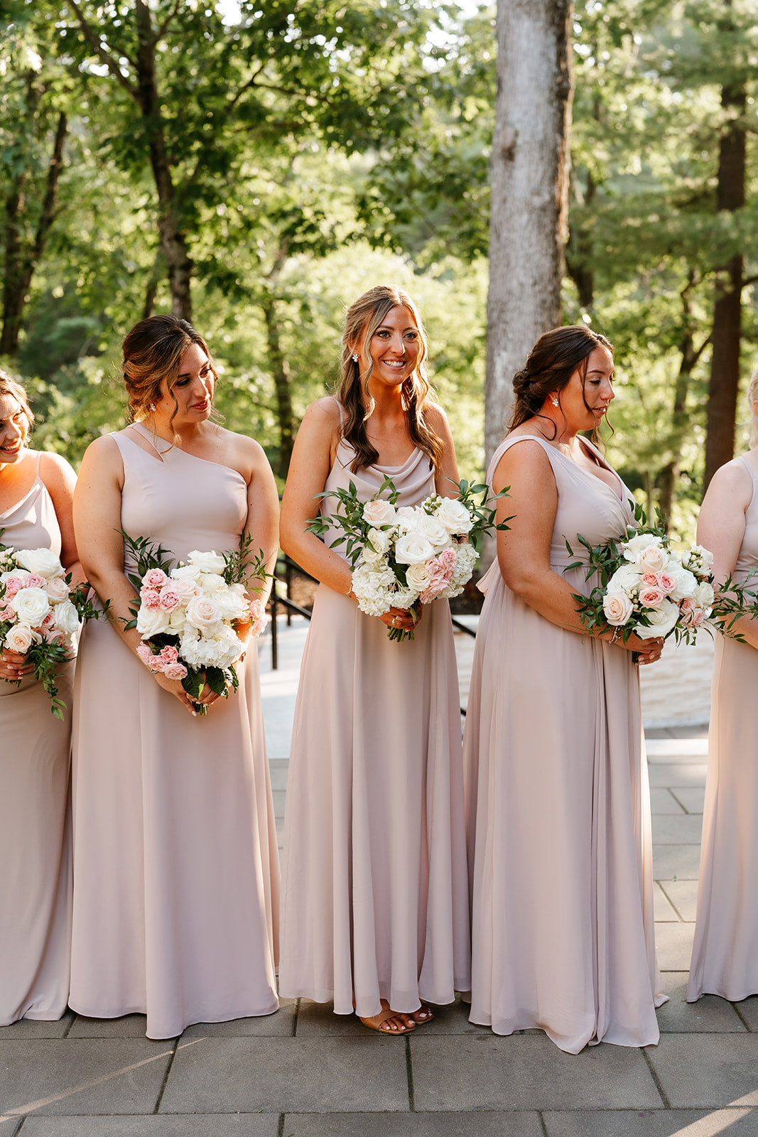 Three bridesmaids wearing matching long, pastel-colored dresses, and holding bouquets of white and pink flowers, stand in a row outdoors with trees in the background.