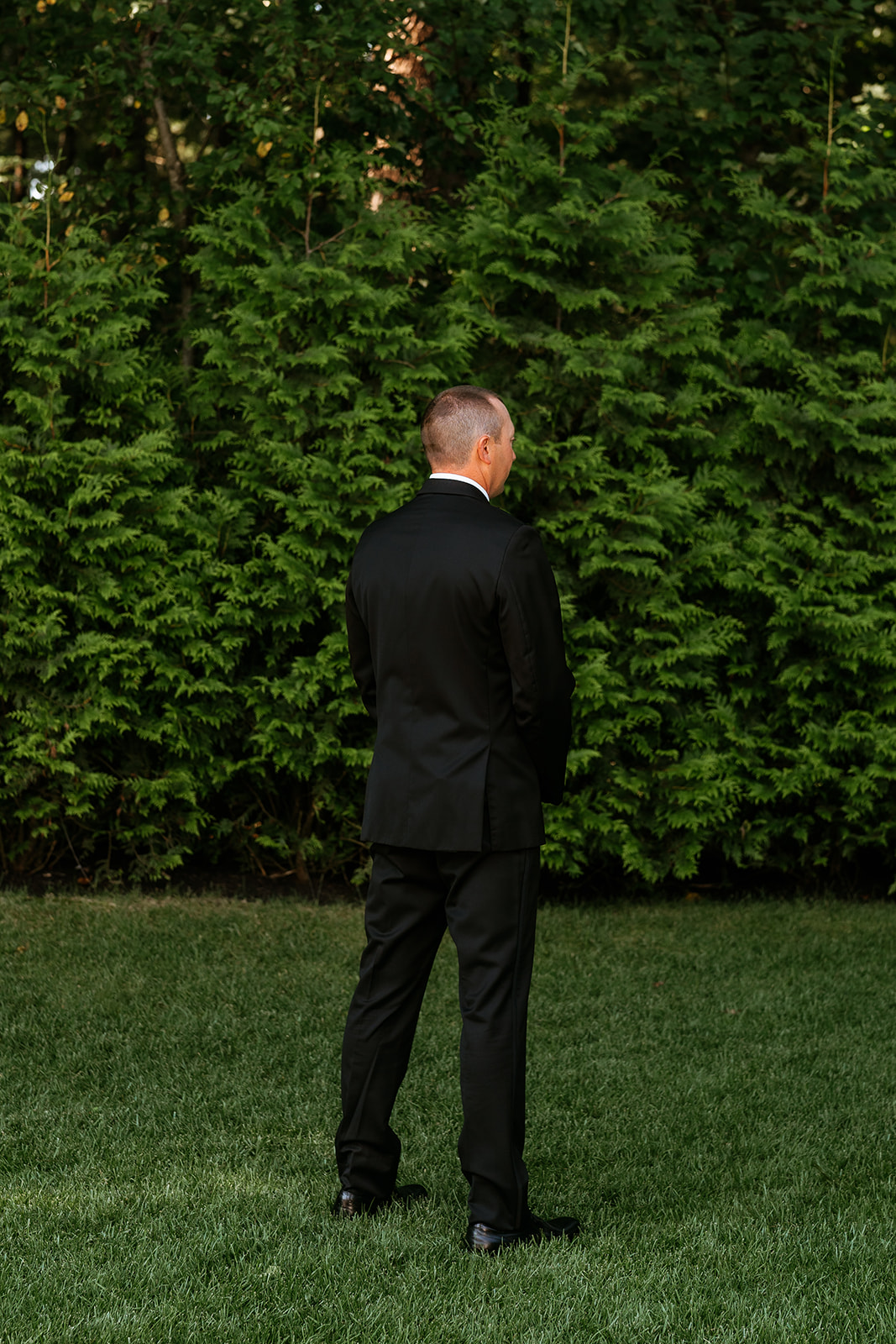 A groom in black suit standing with his back to the camera facing a dense green background waiting to see his bride during a first look.