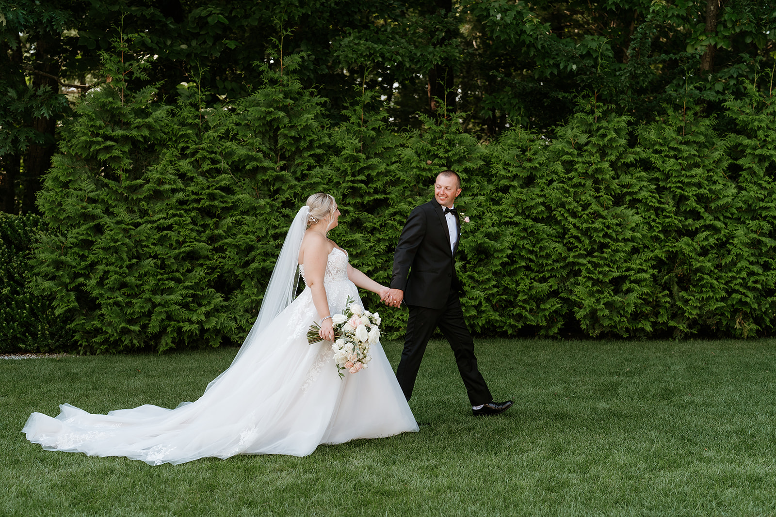 Bride and groom holding hands and walking across a lawn with a backdrop of lush greenery at Lakeview Pavilion