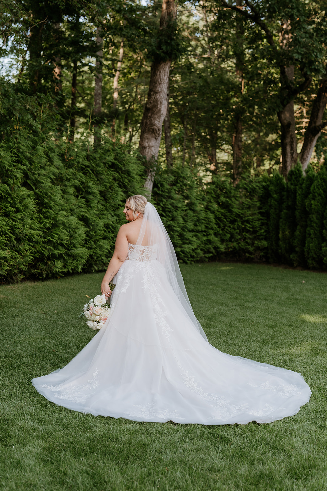 A bride in a white wedding dress with a lace bodice and a full skirt is standing on a grassy lawn with a bouquet in her hands, smiling off to the side. the photograph is in.