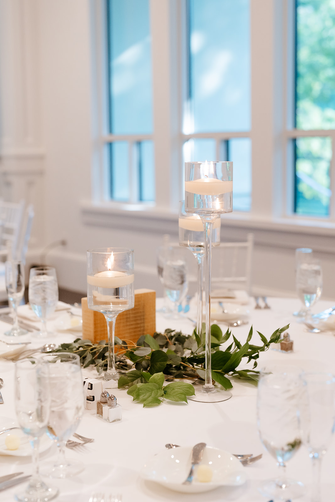 An elegantly set table featuring white linens, tall glass candle holders with lit candles, green foliage decorations, and place settings with silverware and glassware.