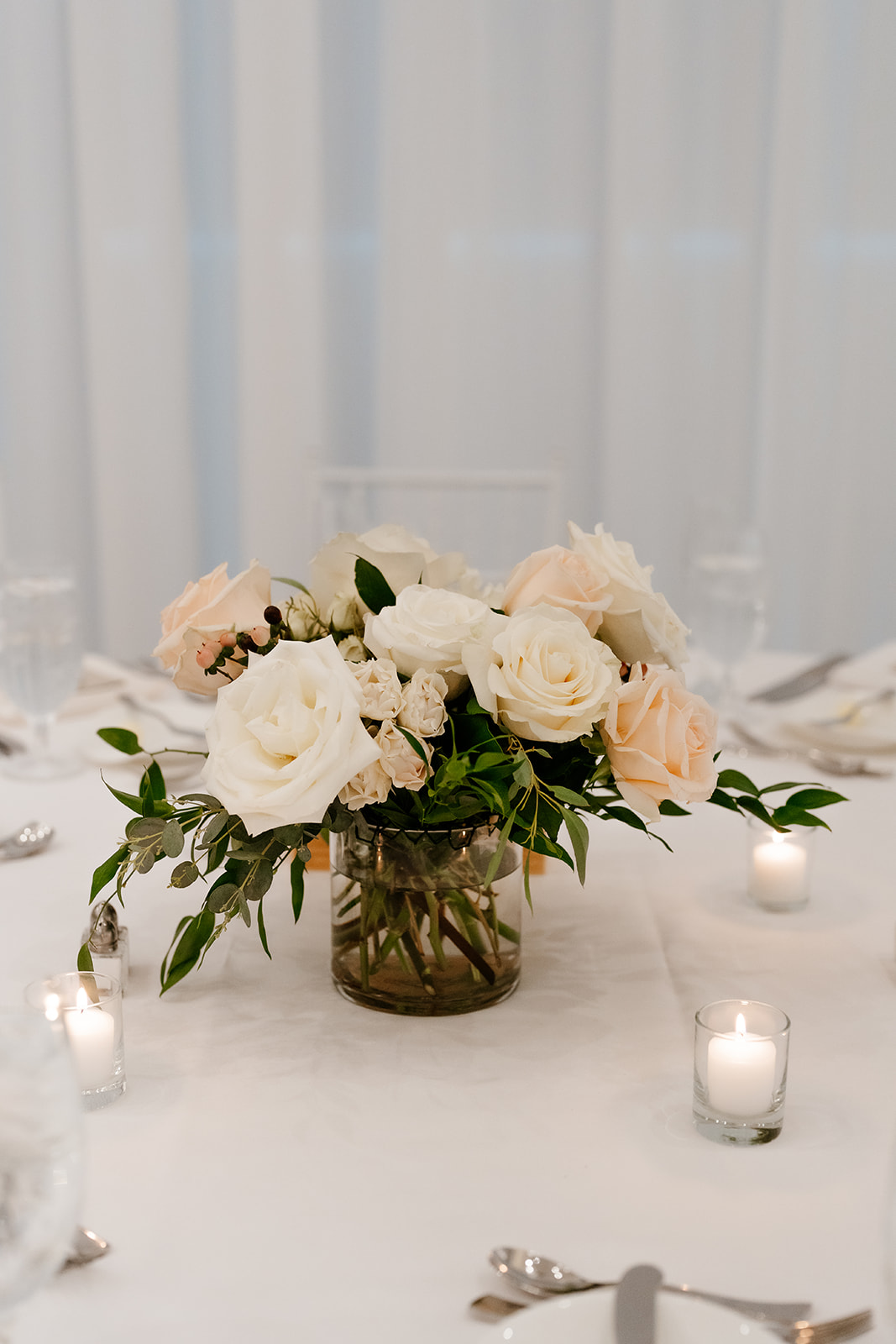 A centerpiece of white and pale pink roses with green foliage in a clear glass vase, accompanied by small lit candles, set upon a white tablecloth.