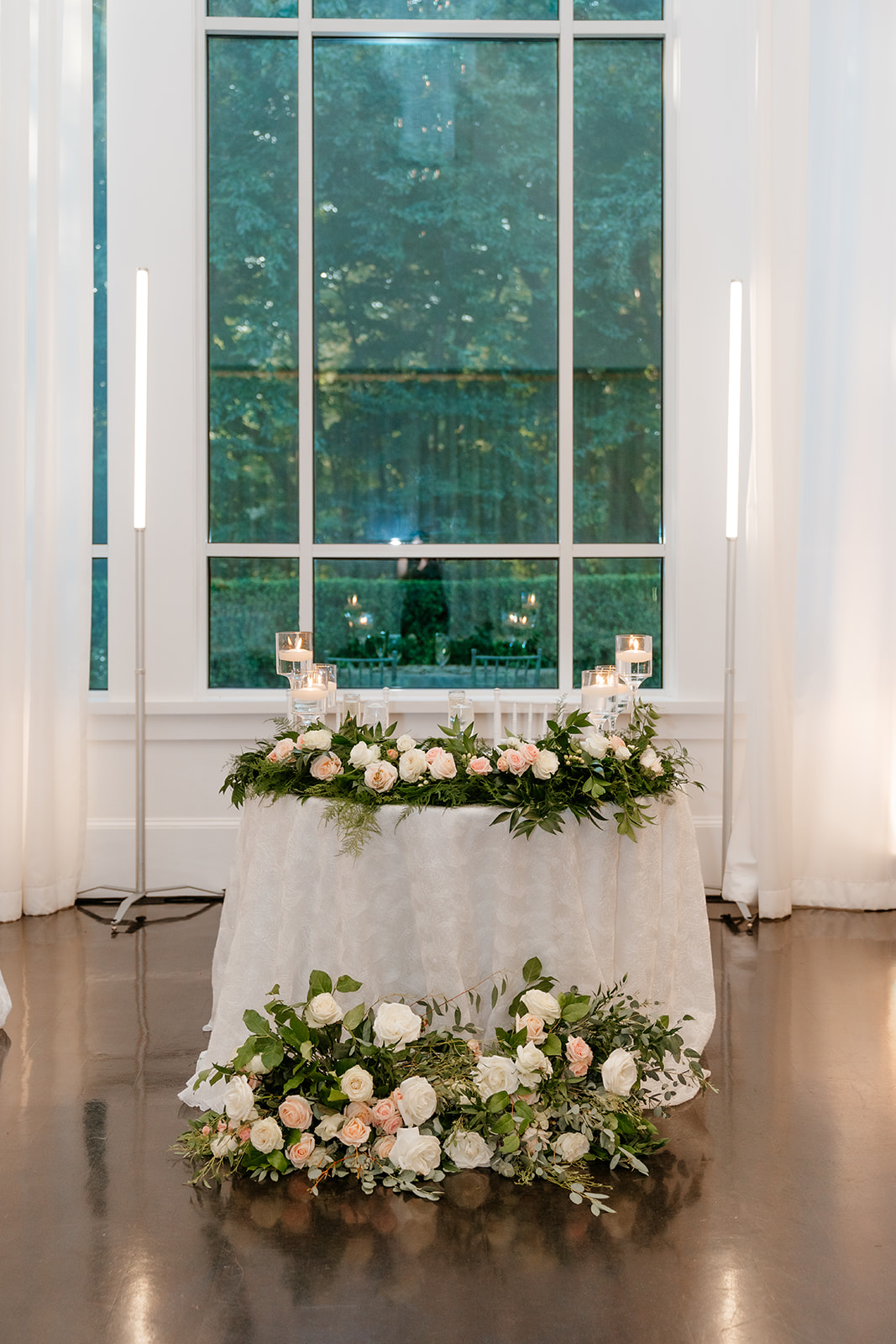 A decorated table with a floral arrangement and candles in front of a large window with white drapes.