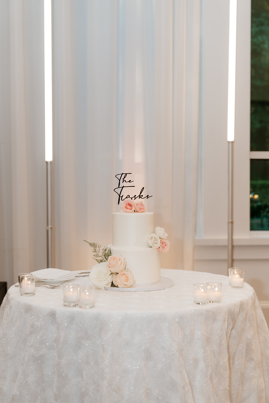 A two-tiered wedding cake with "the trasks" topper, surrounded by lit candles on a table with a sparkly tablecloth, in a room with white curtains and vertical lights.
