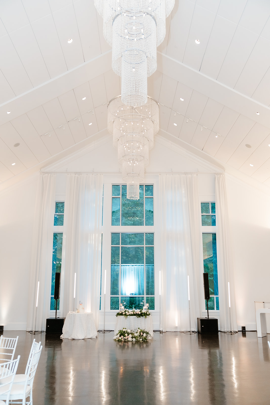 Elegant wedding venue Lakeview Pavilion with a long modern chandelier, high ceilings, white chairs, and floral decorations in front of tall windows.