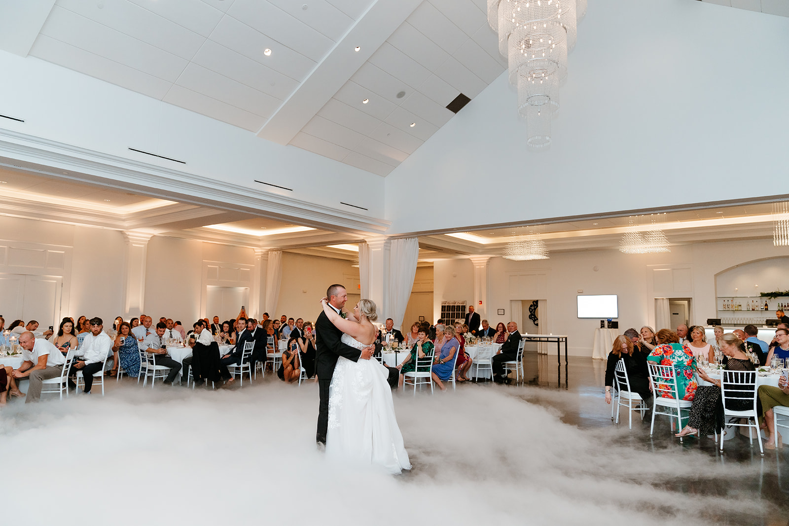 A couple shares their first dance amidst a cloud of fog on the dance floor while guests watch from their tables in a well-lit, elegant banquet hall.