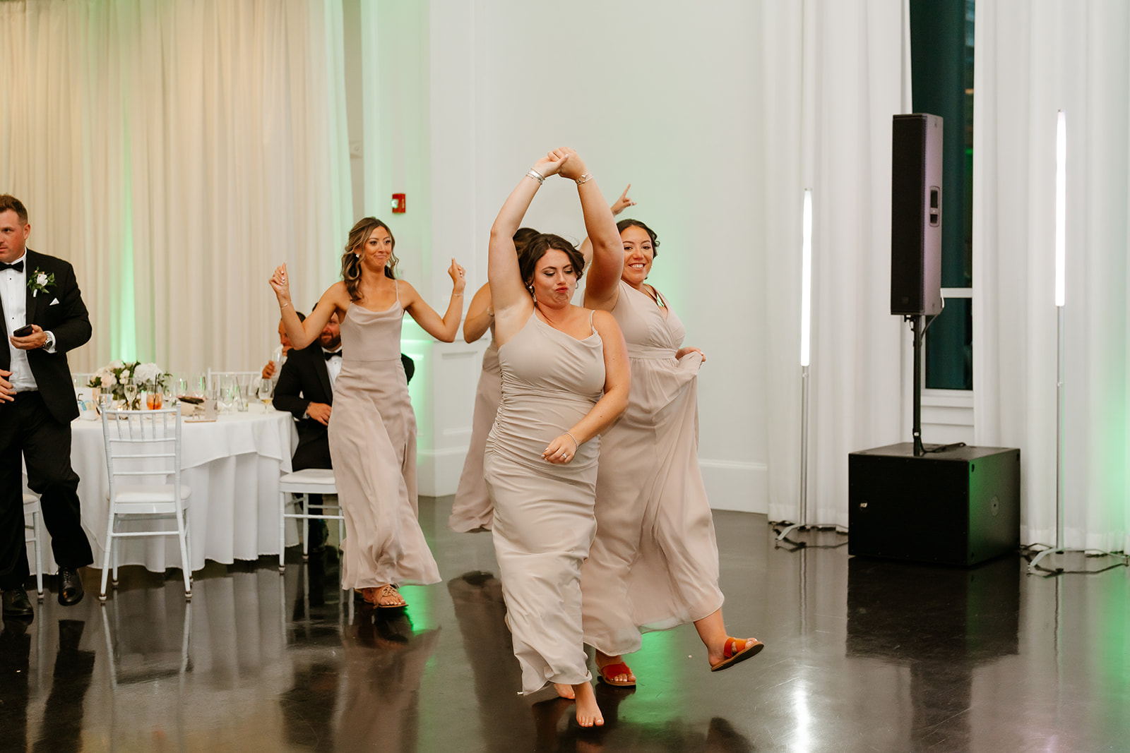 Three women in elegant dresses dancing and having fun at a wedding reception, with a man visible in the background.