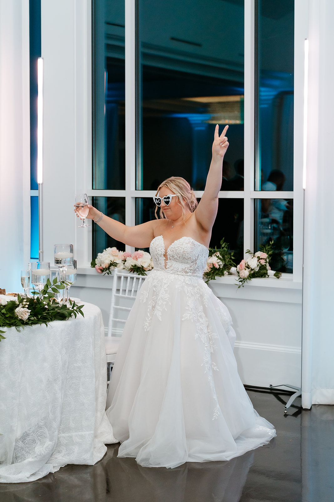 A bride in a white gown with a glass in hand, raising her other hand in a celebratory gesture at a wedding reception.