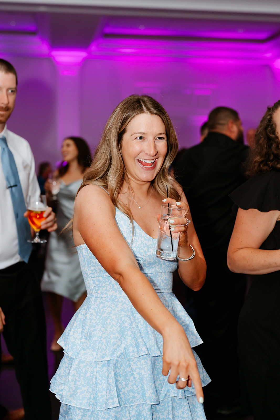 A woman in a blue dress holds a glass of water and smiles at wedding with purple lighting in the background.