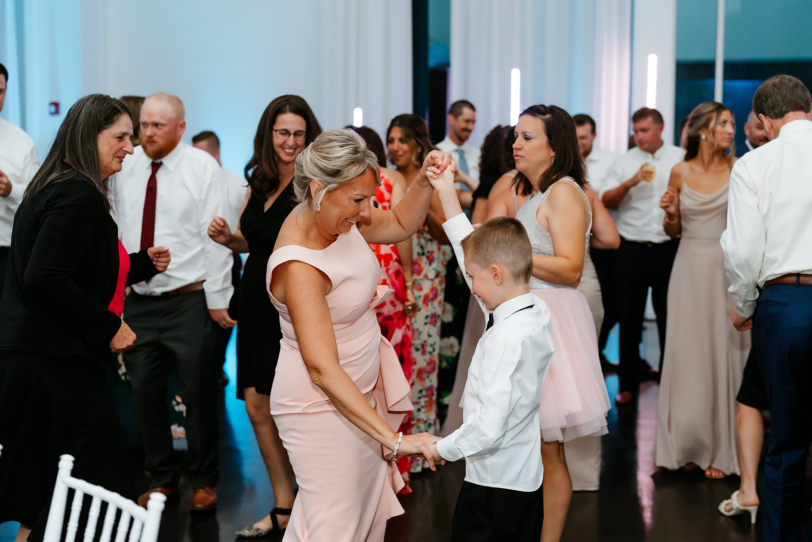 A woman in a pink dress dances with a young boy in a white shirt and black pants at a wedding at Lakeview pavilion while other guests look on and dance in the background.