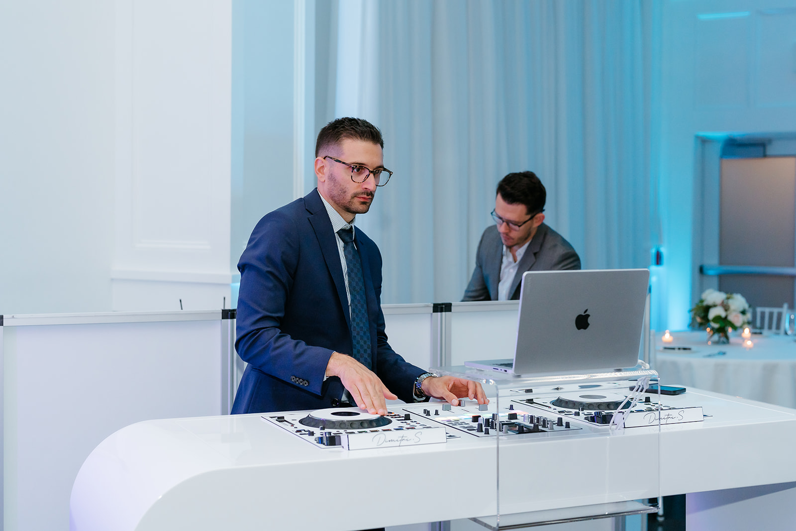 A man in a suit operating dj equipment at an event, with another man in the background working on a laptop.
