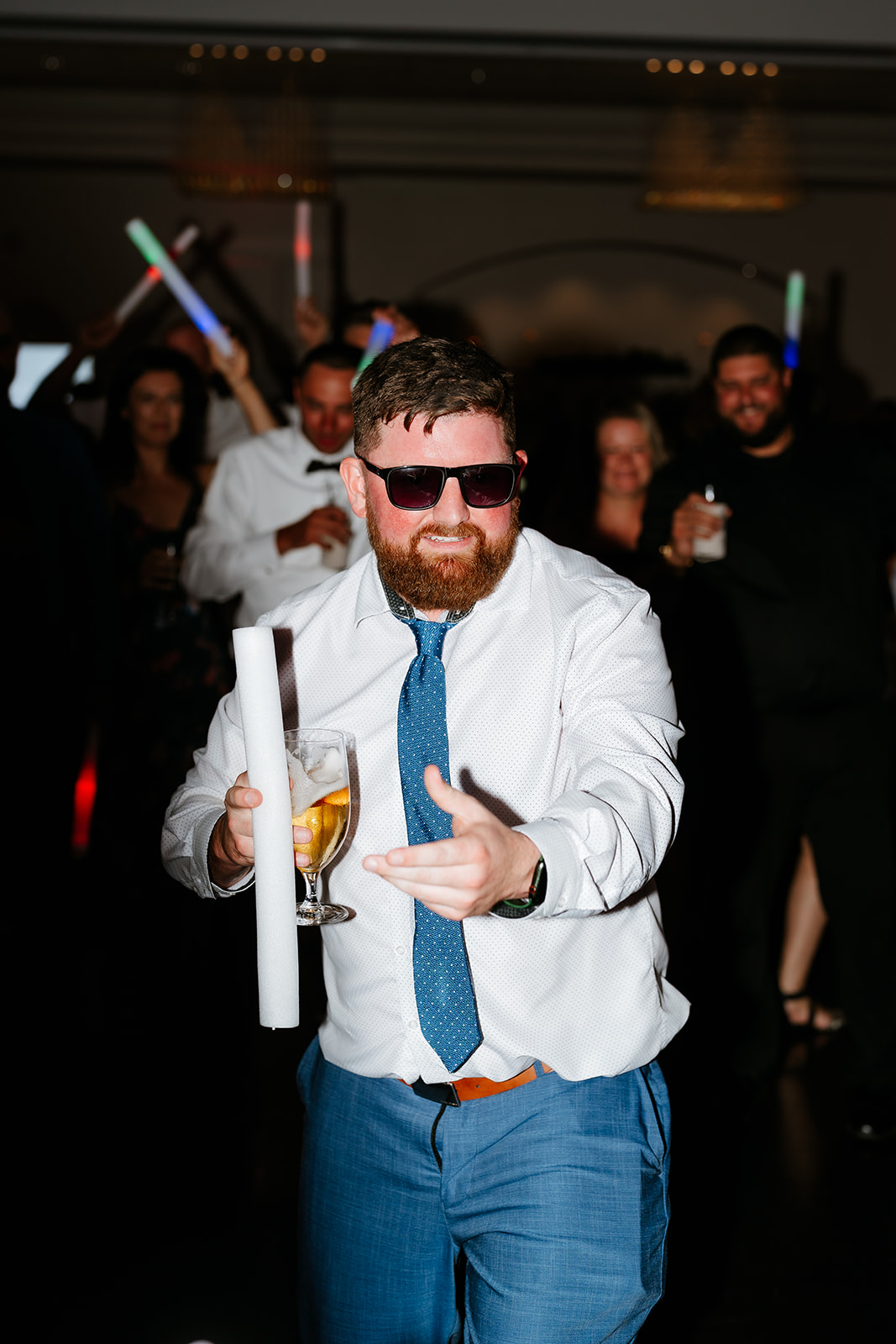 A man in a white shirt and tie, sporting sunglasses and a beard, dances with a drink in hand at a lively wedding with other guests holding colorful glow sticks in the background.