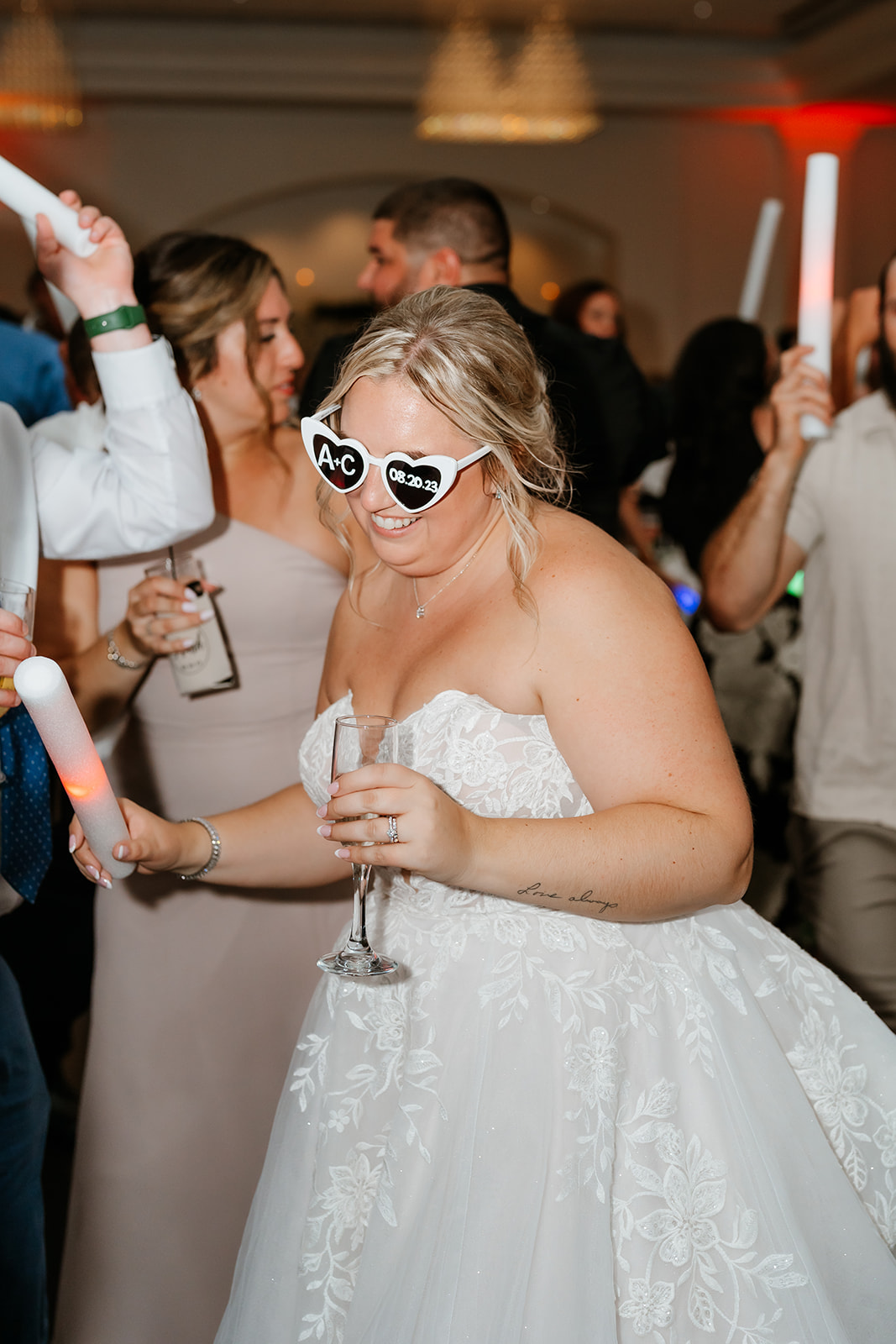 A bride wearing glasses with "bride" written on them is holding a champagne glass and dancing at a wedding reception.