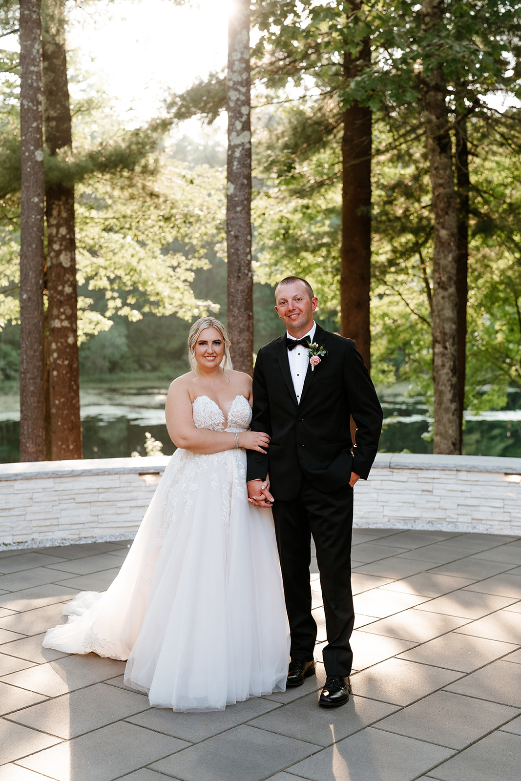 A bride in a white strapless gown and a groom in a black tuxedo with a bow tie are standing together, smiling at the camera, in an outdoor setting with trees and a water feature.