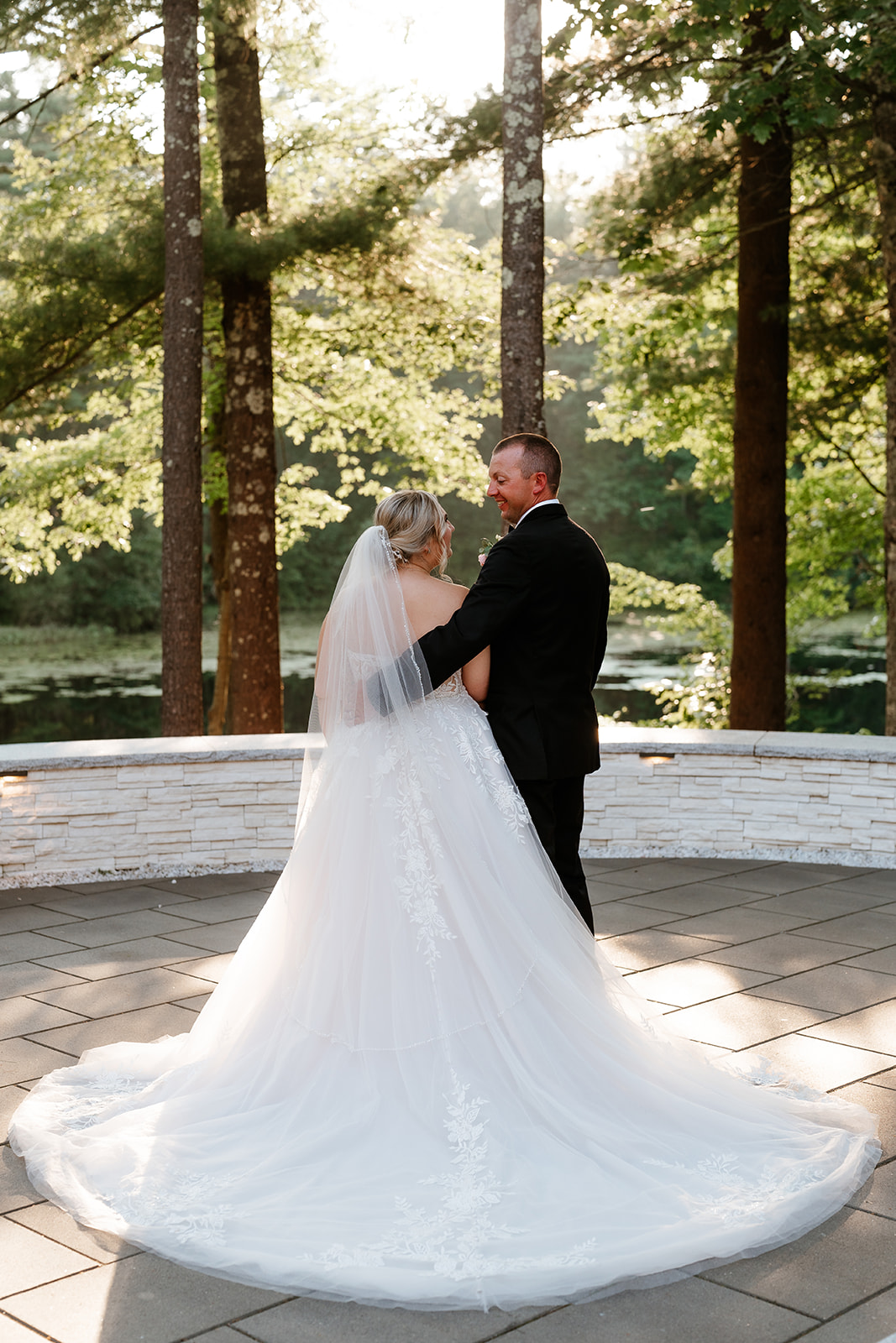 A bride in a white strapless gown and a groom in a black tuxedo with a bow tie are standing together, smiling at the camera, in an outdoor setting with trees and a water feature.