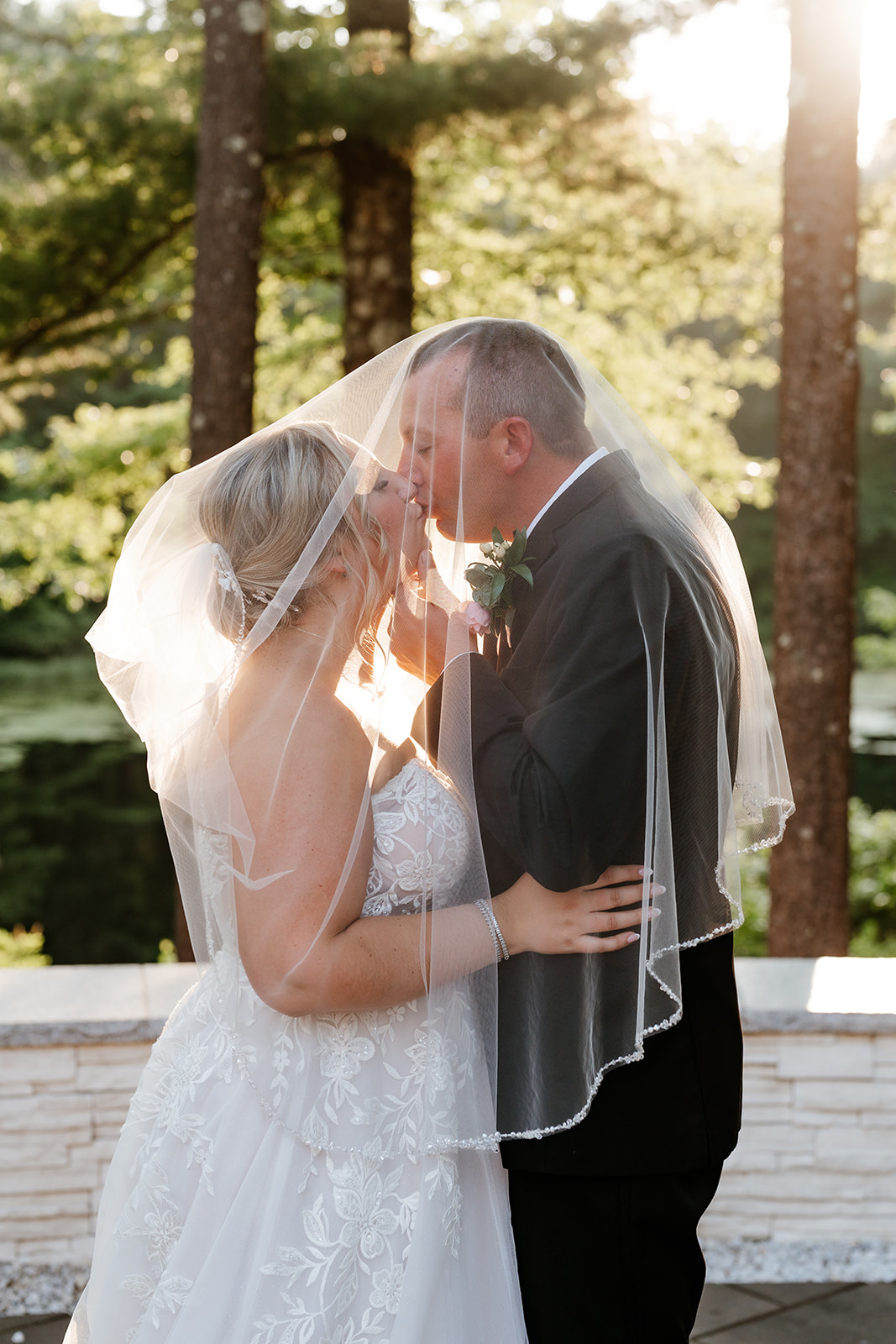 A bride and groom embrace on a sunlit deck surrounded by trees, while kissing