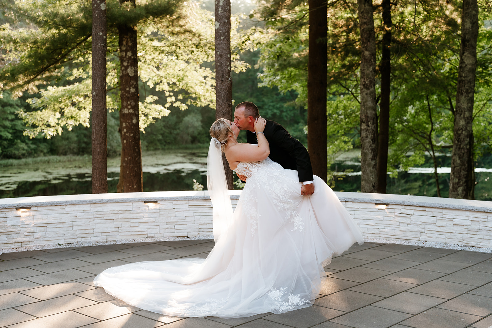 A bride and groom embrace on a sunlit deck surrounded by trees, while kissing