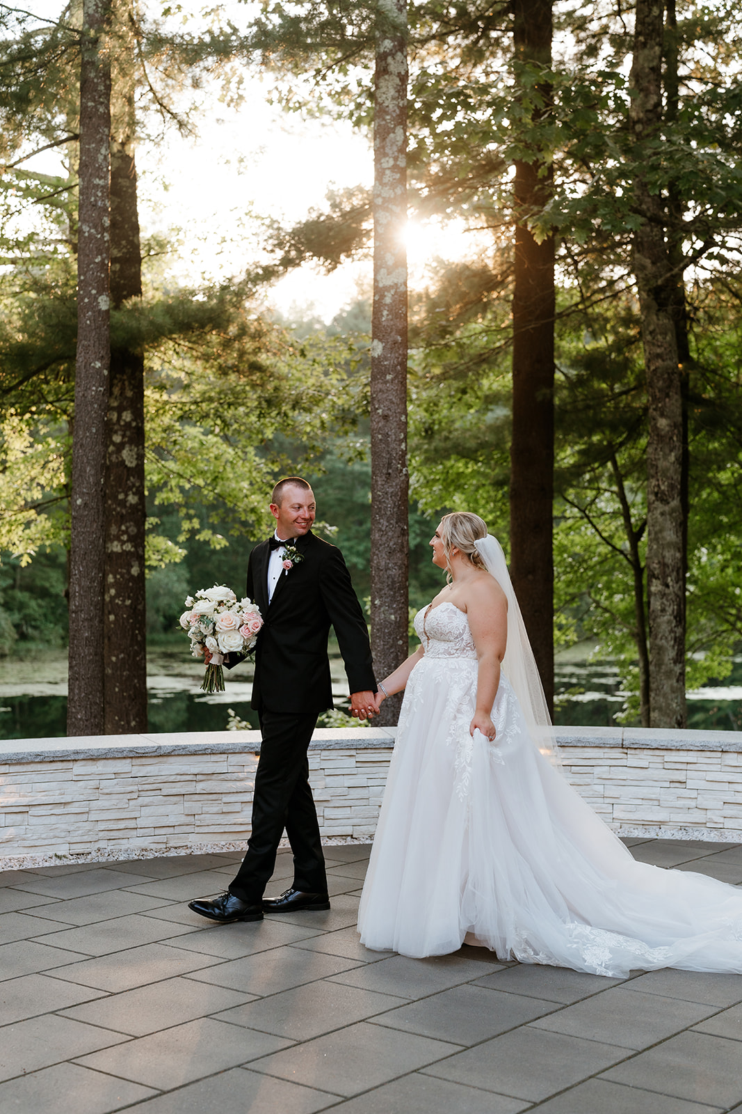 A couple dressed in wedding attire holding hands and looking at each other, with trees and a soft sunlight background.