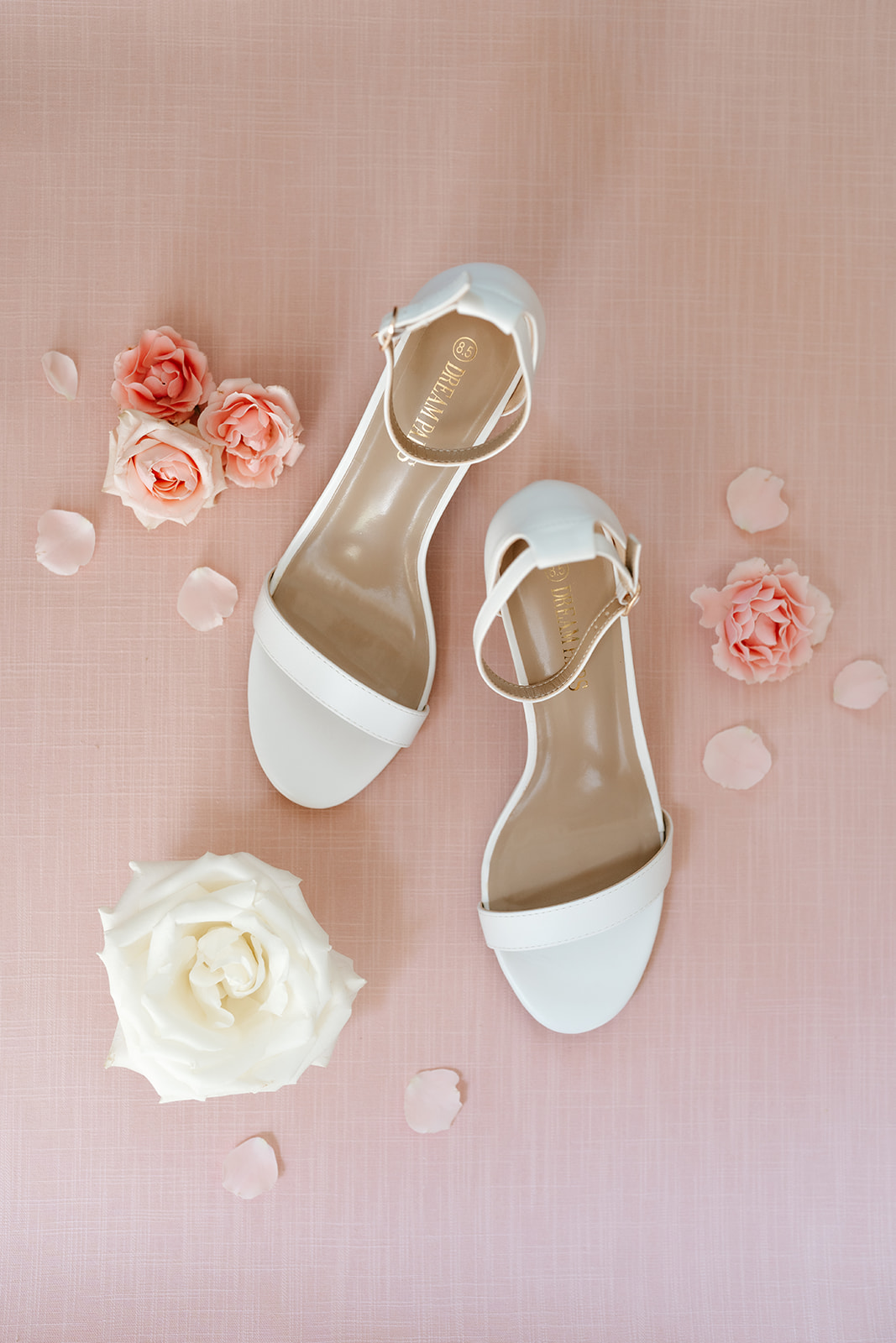A pair of white women's high heel shoes arranged neatly on a pink background, surrounded by scattered rose petals and a few full blooms.