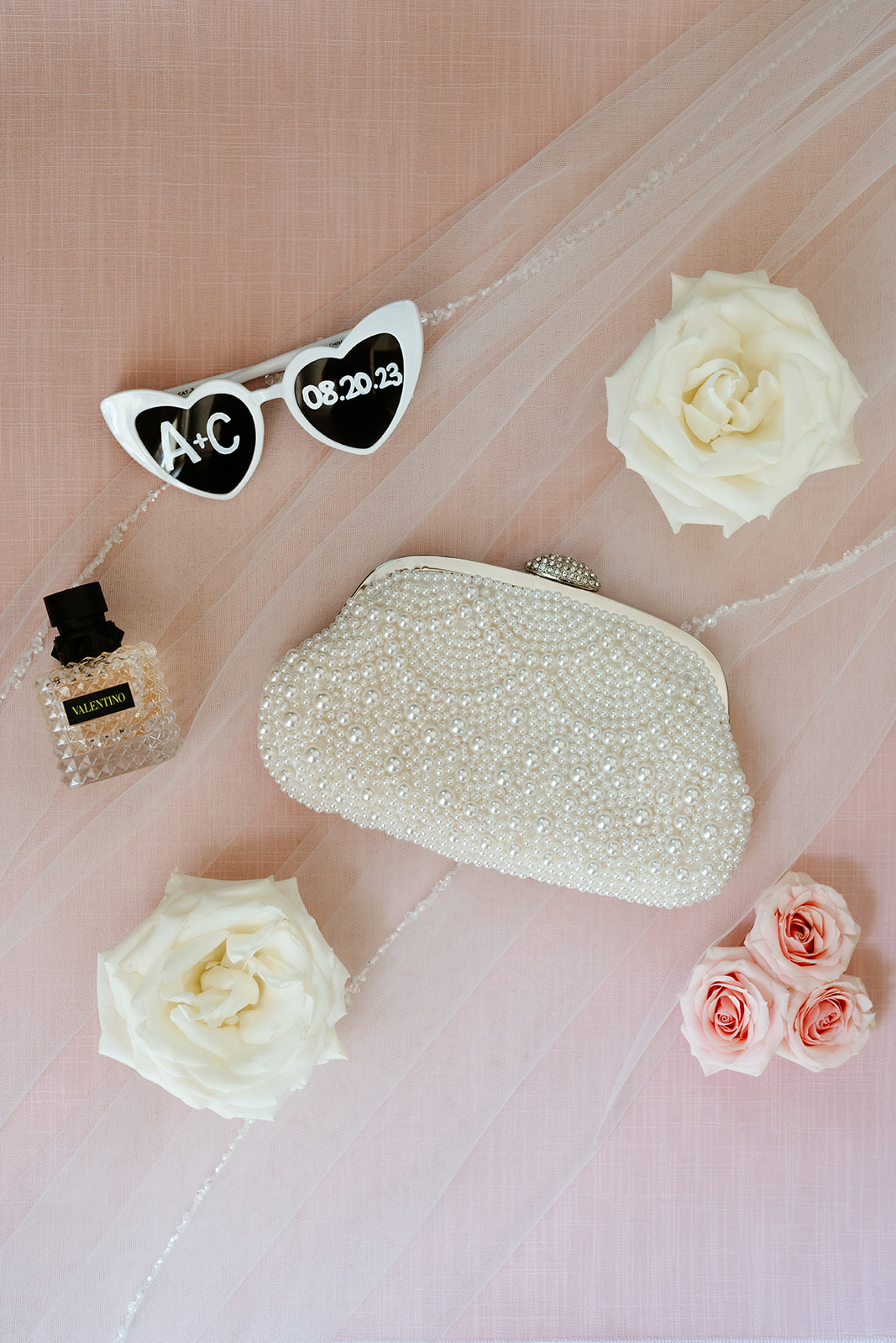 A pair of white women's sunglasses, perfume and handbag surrounded by scattered rose petals and a few full blooms.