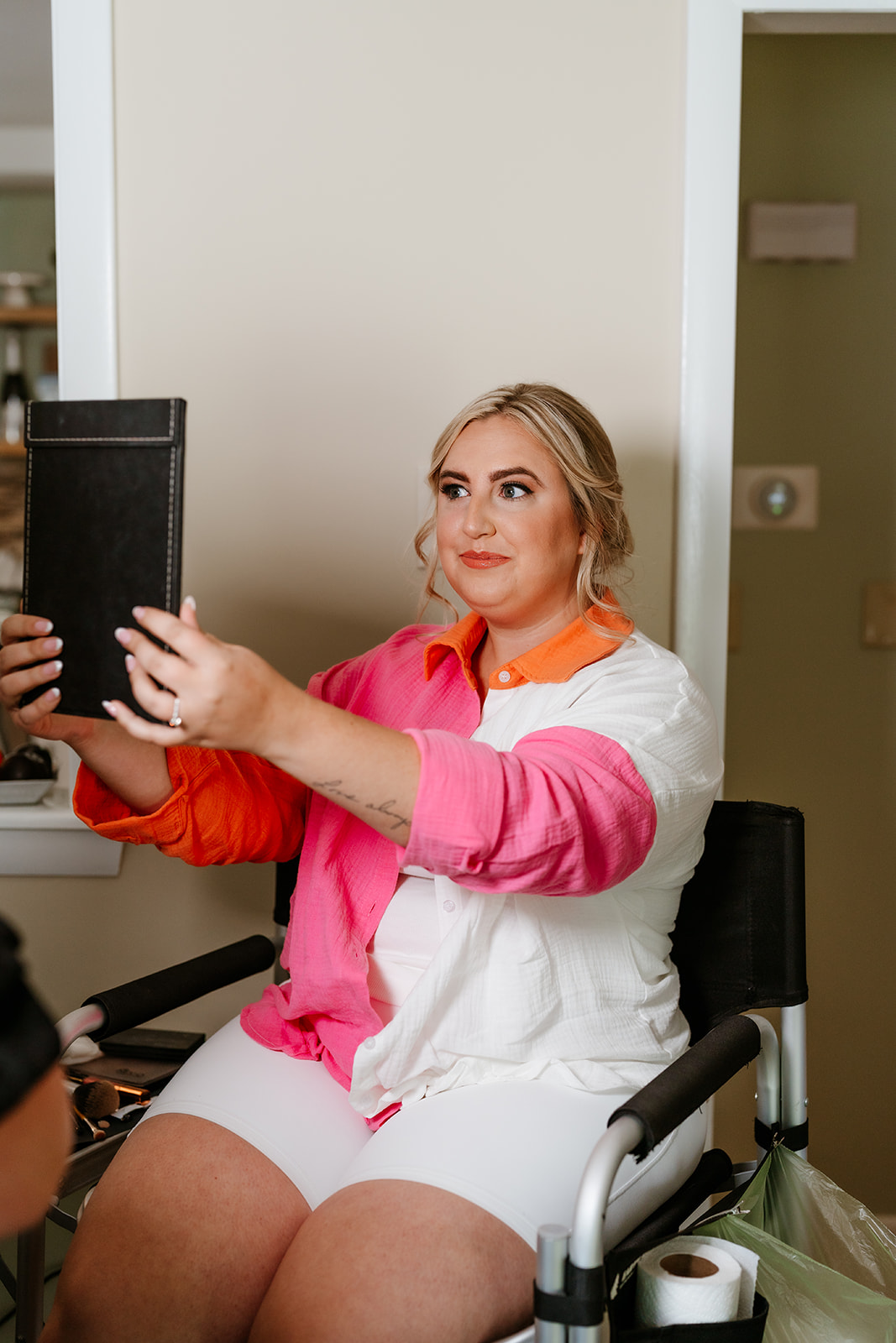 A woman in a white chair looks at a mirror she is holding, wearing a white shirt with pink detailing and a bright orange collar, with her hair styled up.