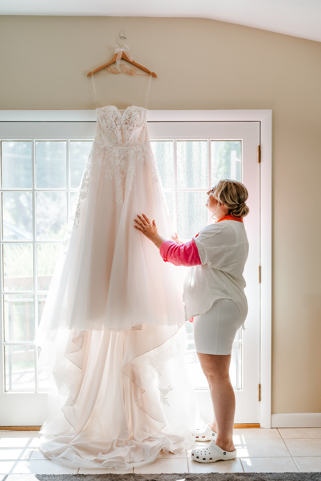 A woman in a white robe stands on tiptoe and reaches out to touch a white wedding dress hanging in front of a window in a well-lit room.