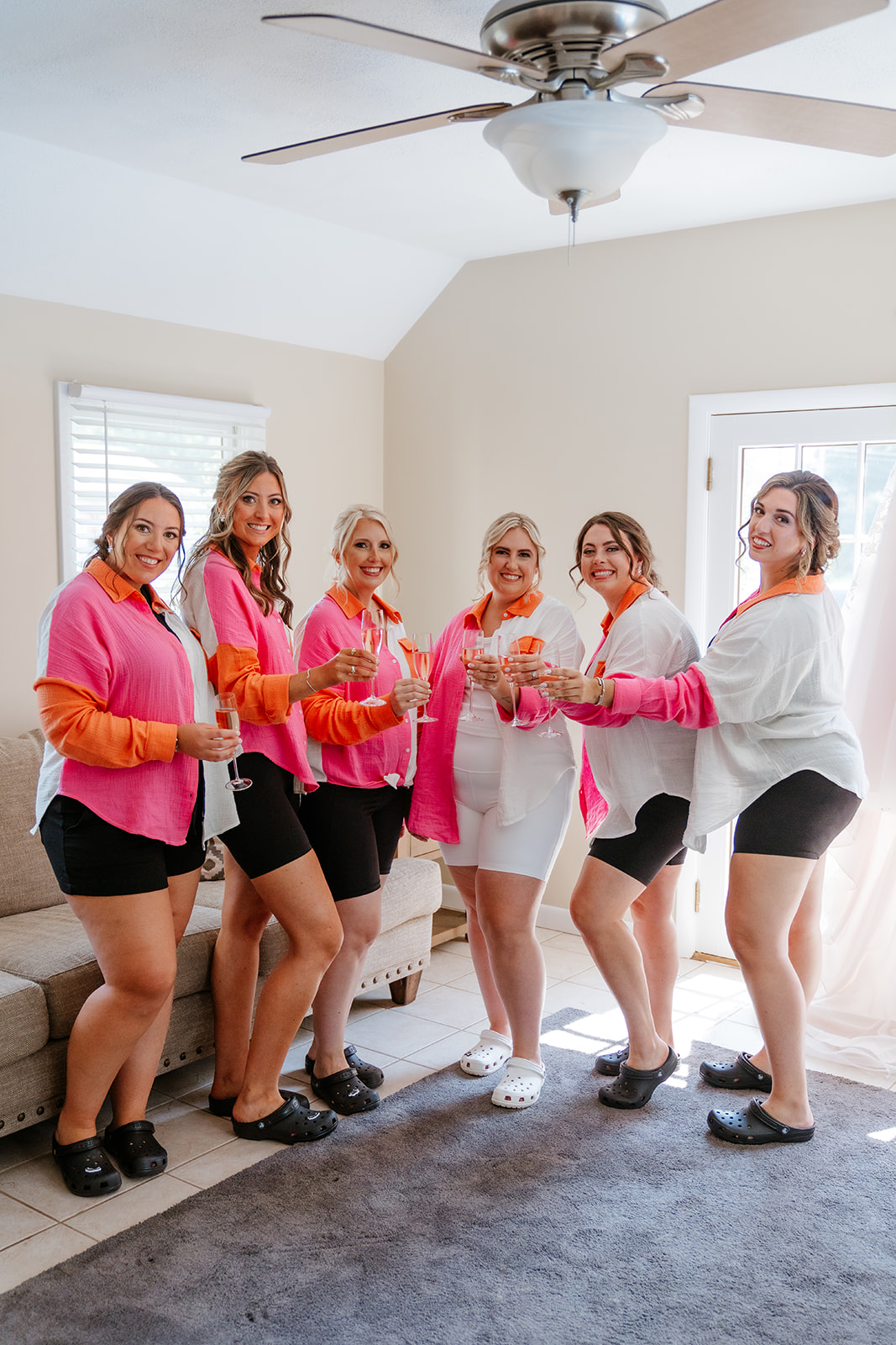 Bride and bridesmaids in white shorts and pink buttoned shirts, holding champagne are posing for a photo in their getting ready outfits for bride's wedding at lakeview pavilion