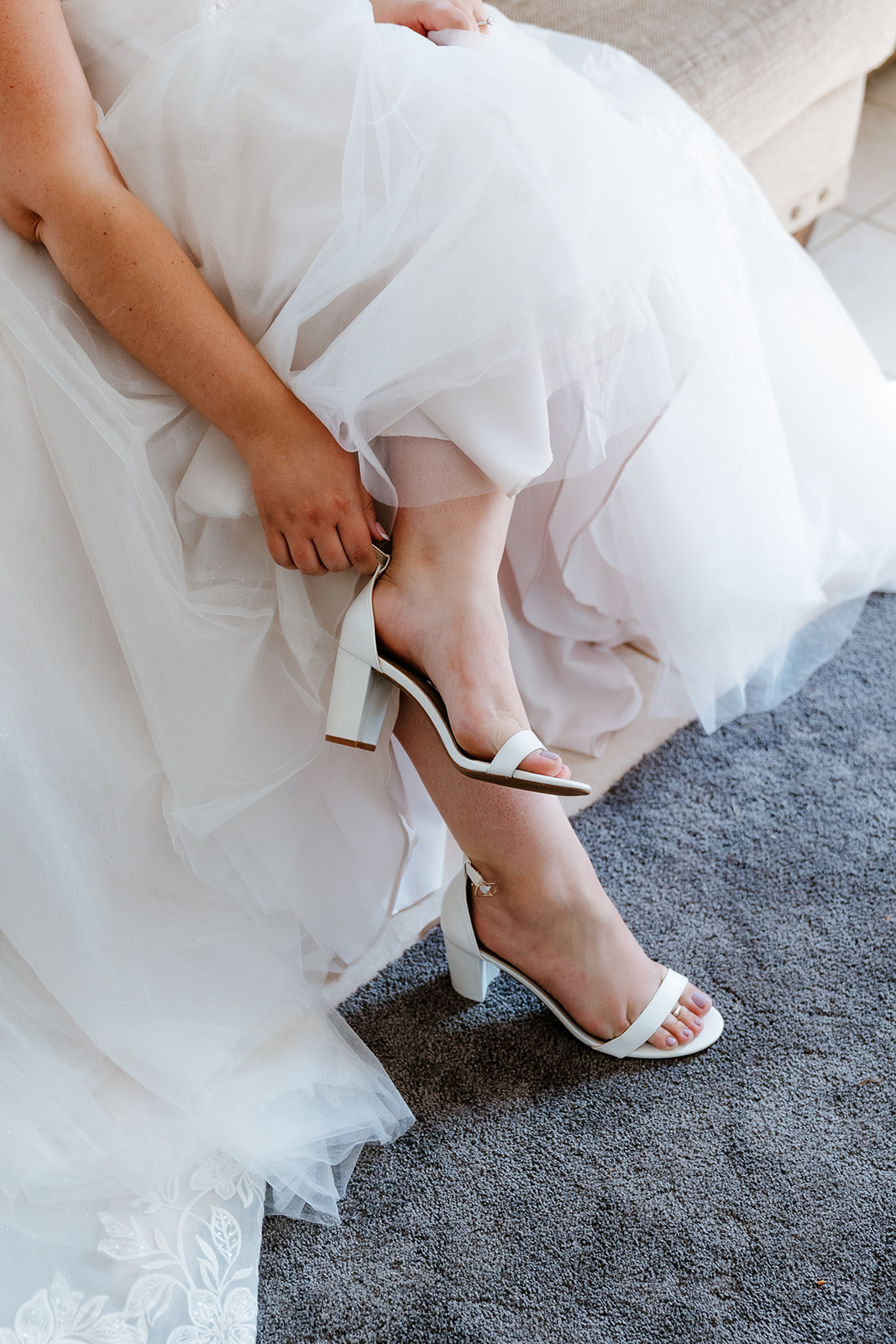 A bride is seated while adjusting the strap on her white heeled sandal beneath her layered tulle wedding dress.