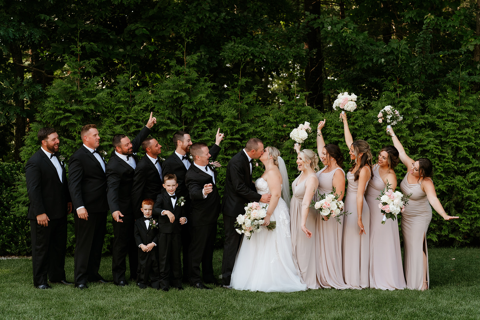 A wedding party posing outdoors, with a groom kissing the bride while the rest of the group, dressed in formal attire, engages in various playful gestures.