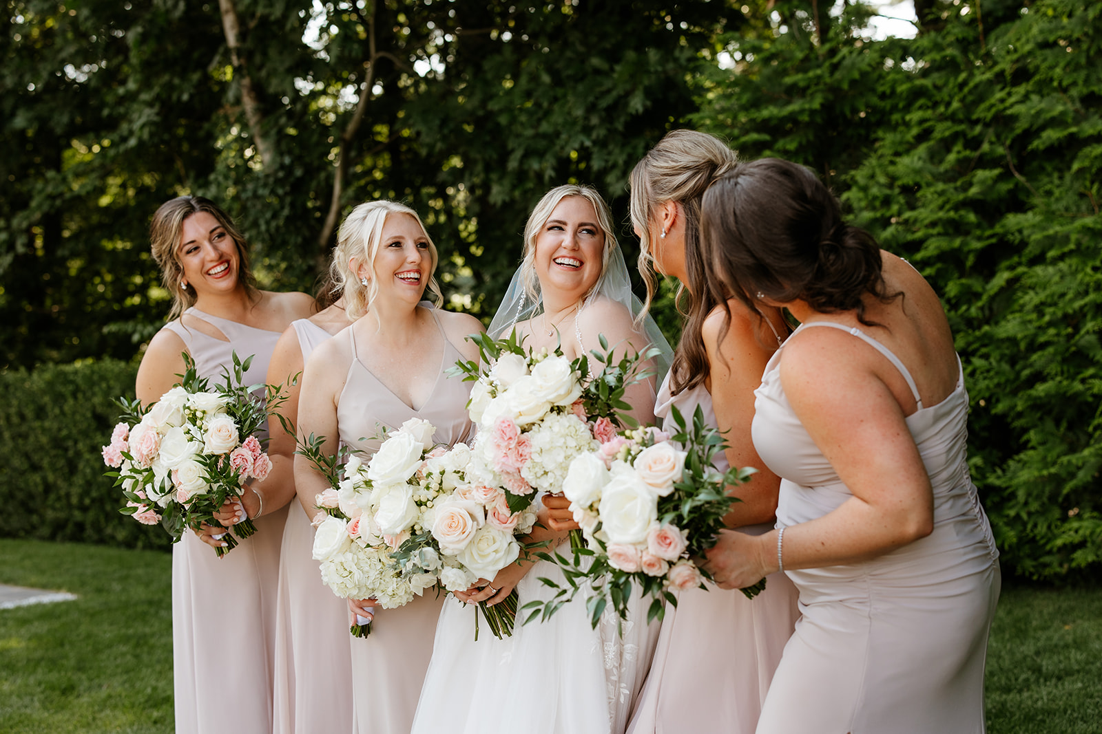 Bridesmaids coordinating dresses pose with a bride, holding bouquets against a backdrop of greenery at Lakeview Pavilion