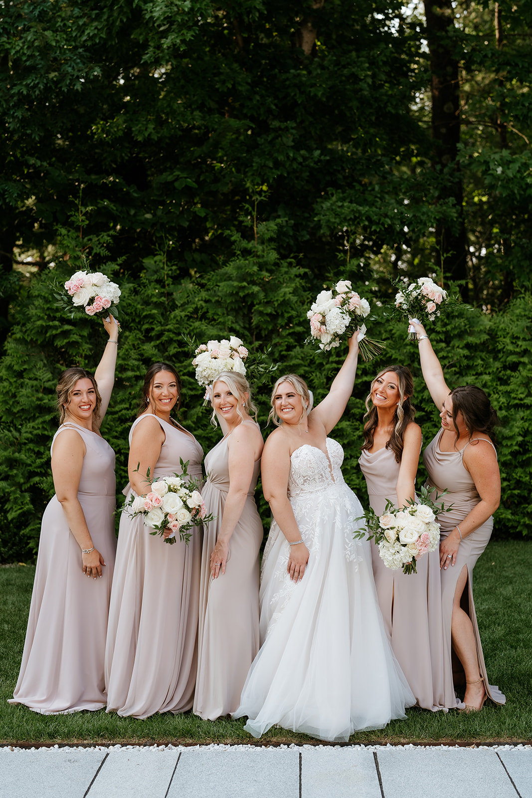 A bride in a white dress stands with four bridesmaids in matching pale pink dresses, all holding bouquets and raising one arm joyfully in front of a green hedge at Lakeview Pavilion