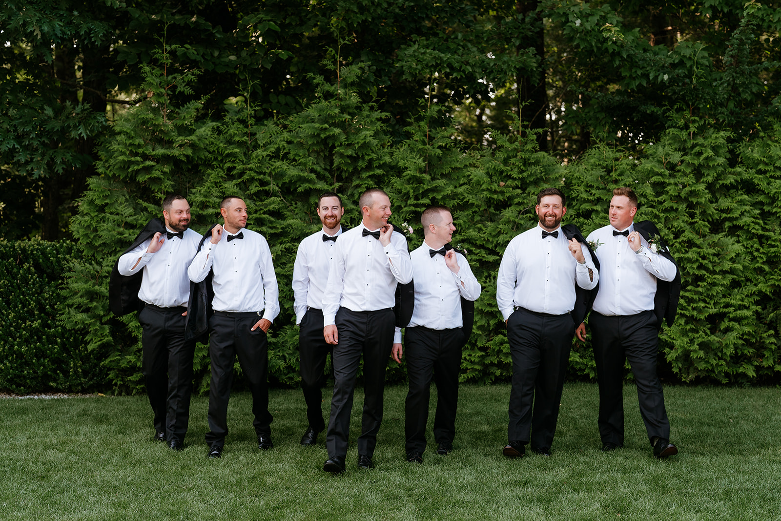 A group of men dressed in black tuxedos with boutonnieres stand in a line on a grassy area with a backdrop of dense green shrubbery at Lakeview Pavilion.