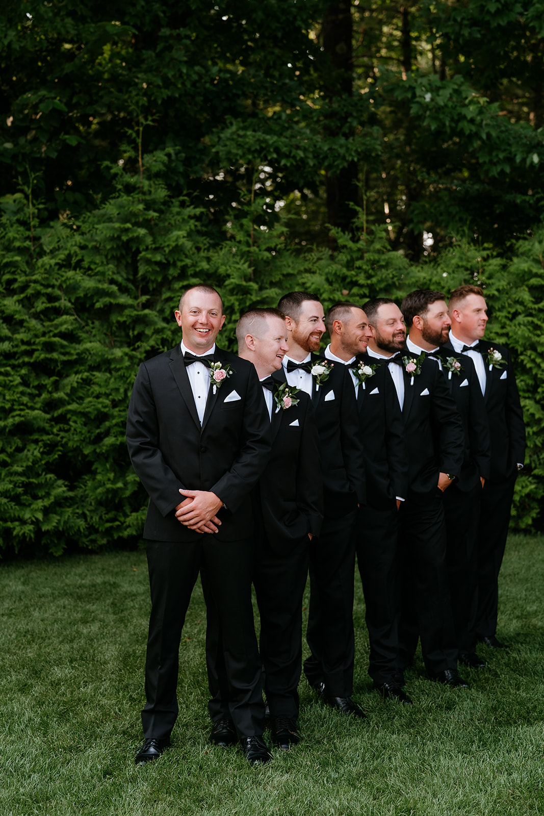 A group of men dressed in black tuxedos with boutonnieres stand in a line on a grassy area with a backdrop of dense green shrubbery at Lakeview Pavilion.