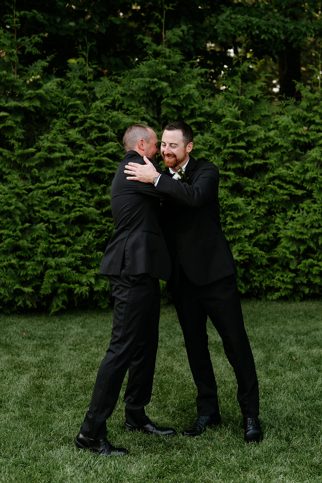 Two men in black suits embracing and smiling in front of a green, leafy backdrop at Lakeview Pavilion.