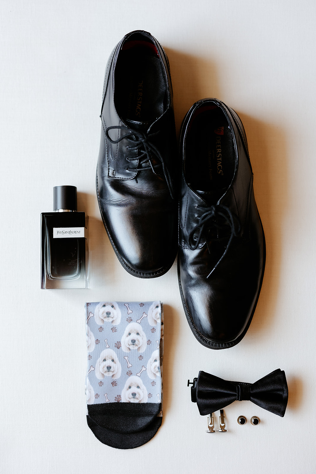 A pair of polished black dress shoes arranged neatly beside a bottle of cologne, patterned socks, and a bow tie with cufflinks set on a light background.