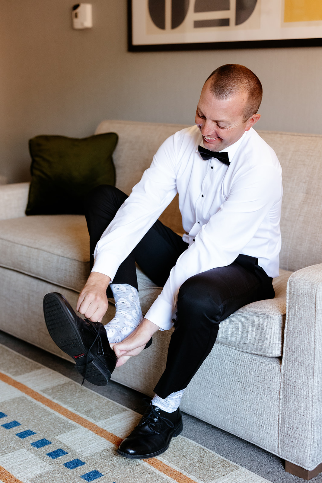 A groom  in a white shirt and black bow tie is sitting on a couch tying his dress shoe.
