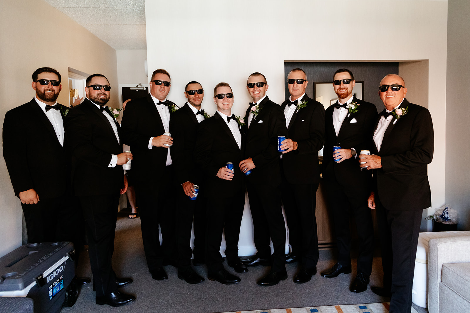Groom and groomsmen in formal attire with sunglasses, holding drinks, pose together in a room.