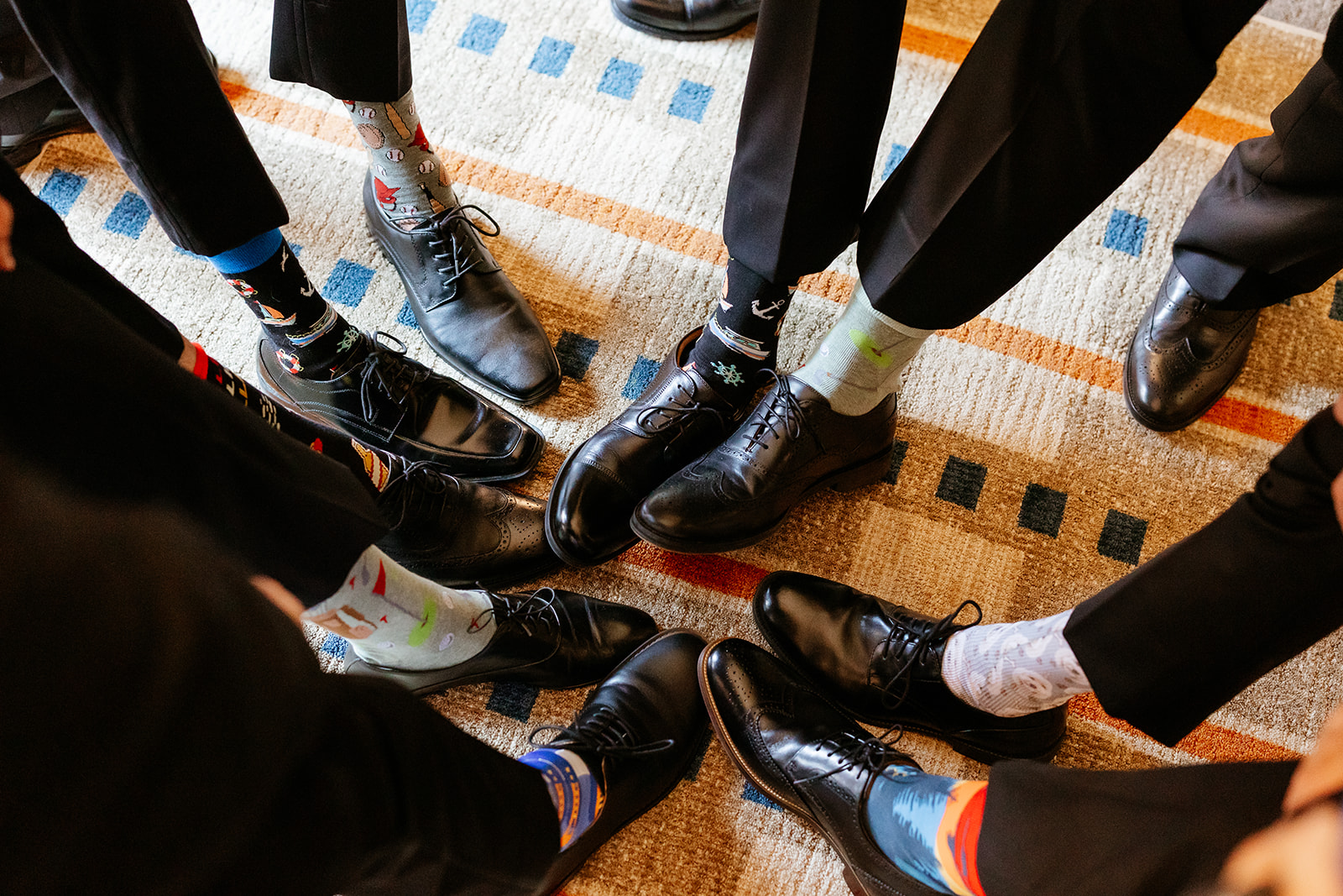 A group of individuals wearing formal black shoes and a variety of colorful, patterned socks stand closely together, showcasing a contrast between traditional formal attire and playful sock choices.
