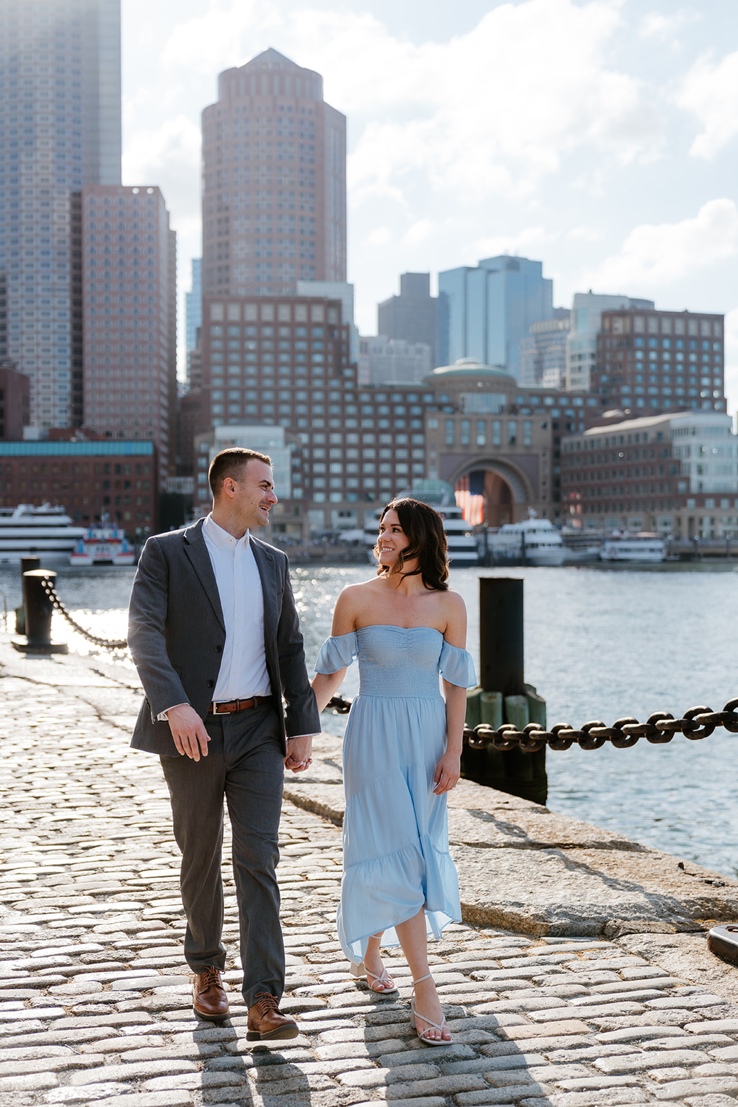 A couple walks hand in hand along a waterfront with city buildings in the background at Fan Pier Park as they take Boston engagement photos