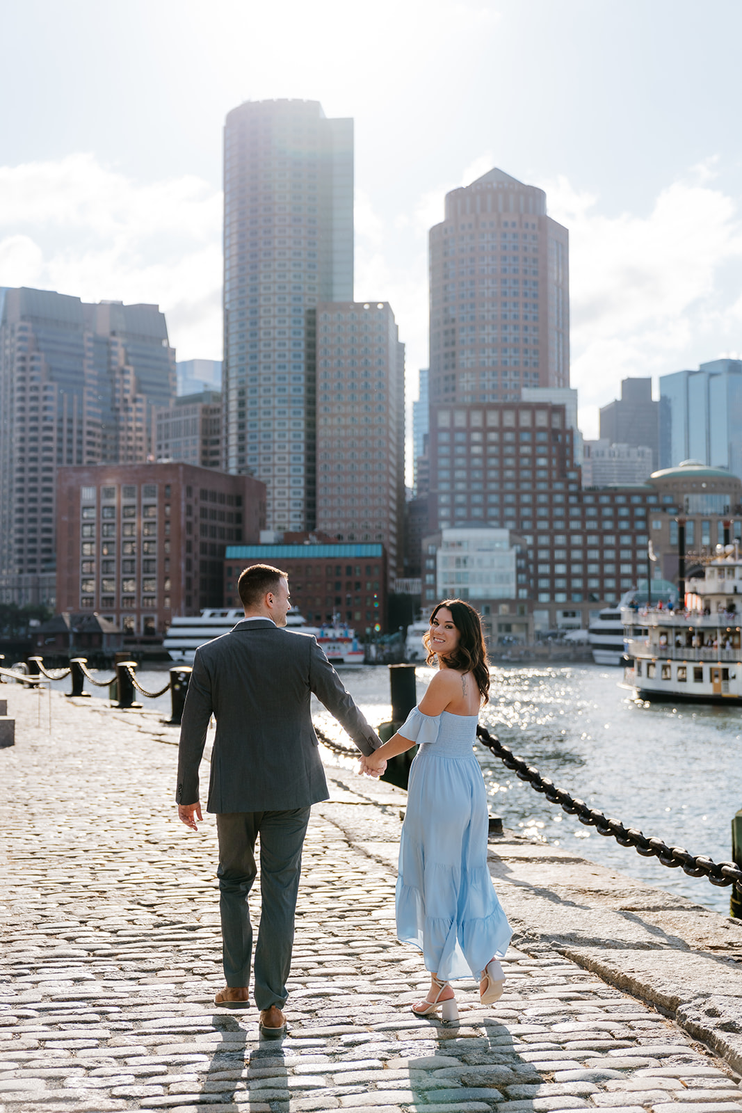 A couple walks hand in hand along a waterfront with city buildings in the background at Fan pier Park as they take Boston engagement photos