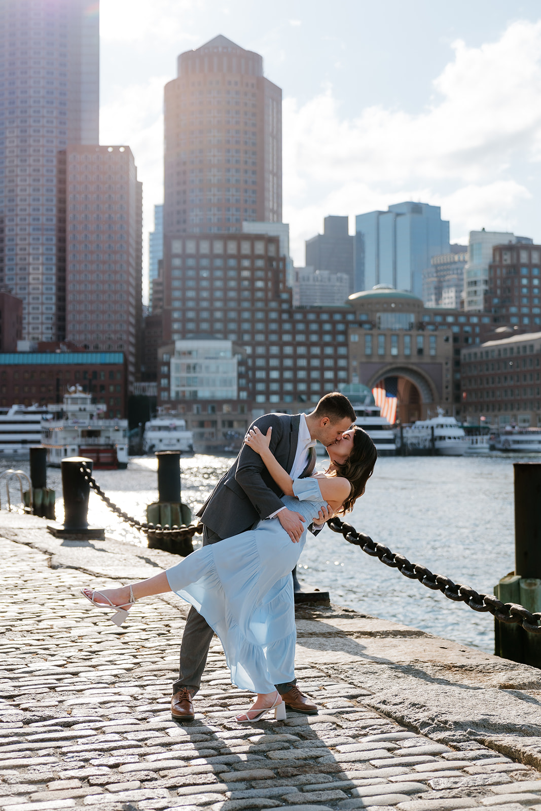 Couple sharing a romantic kiss on a sunny waterfront with city buildings in the background at Fan pier Park as they take Boston engagement photos