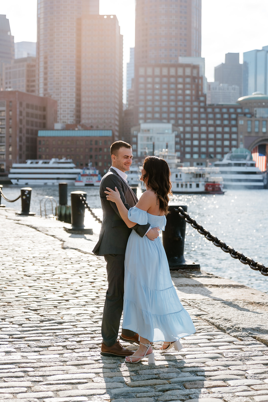Couple on a sunny waterfront with city buildings in the background at Fan pier Park as they take Boston engagement photos
