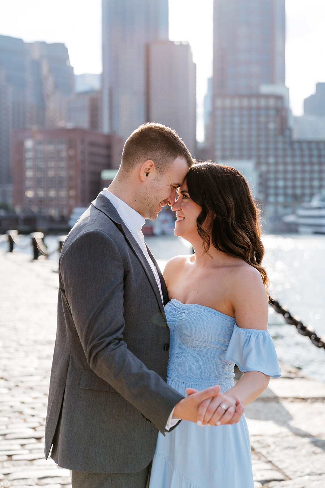 Couple looking at each other while bumping foreheads on a sunny waterfront with city buildings in the background at Fan pier Park as they take Boston engagement photos