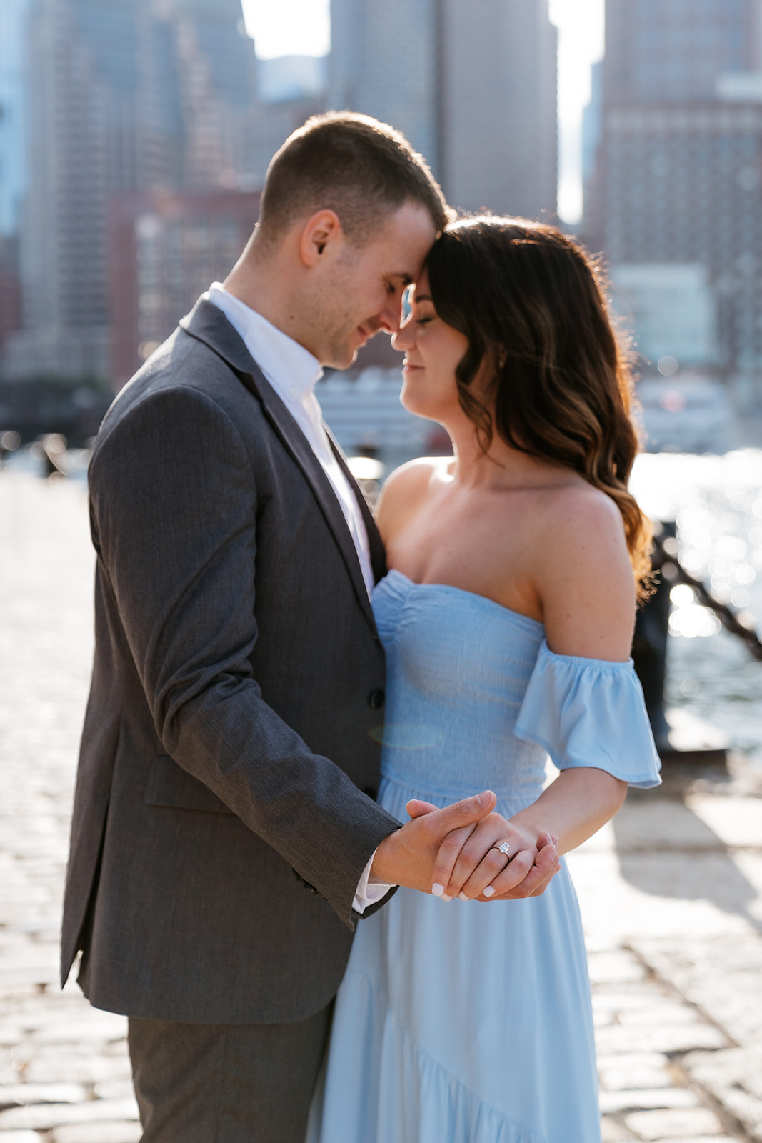 A couple holding hands and sharing an intimate moment by the waterfront with city buildings in the background at fan pier park as they take Boston engagement photos